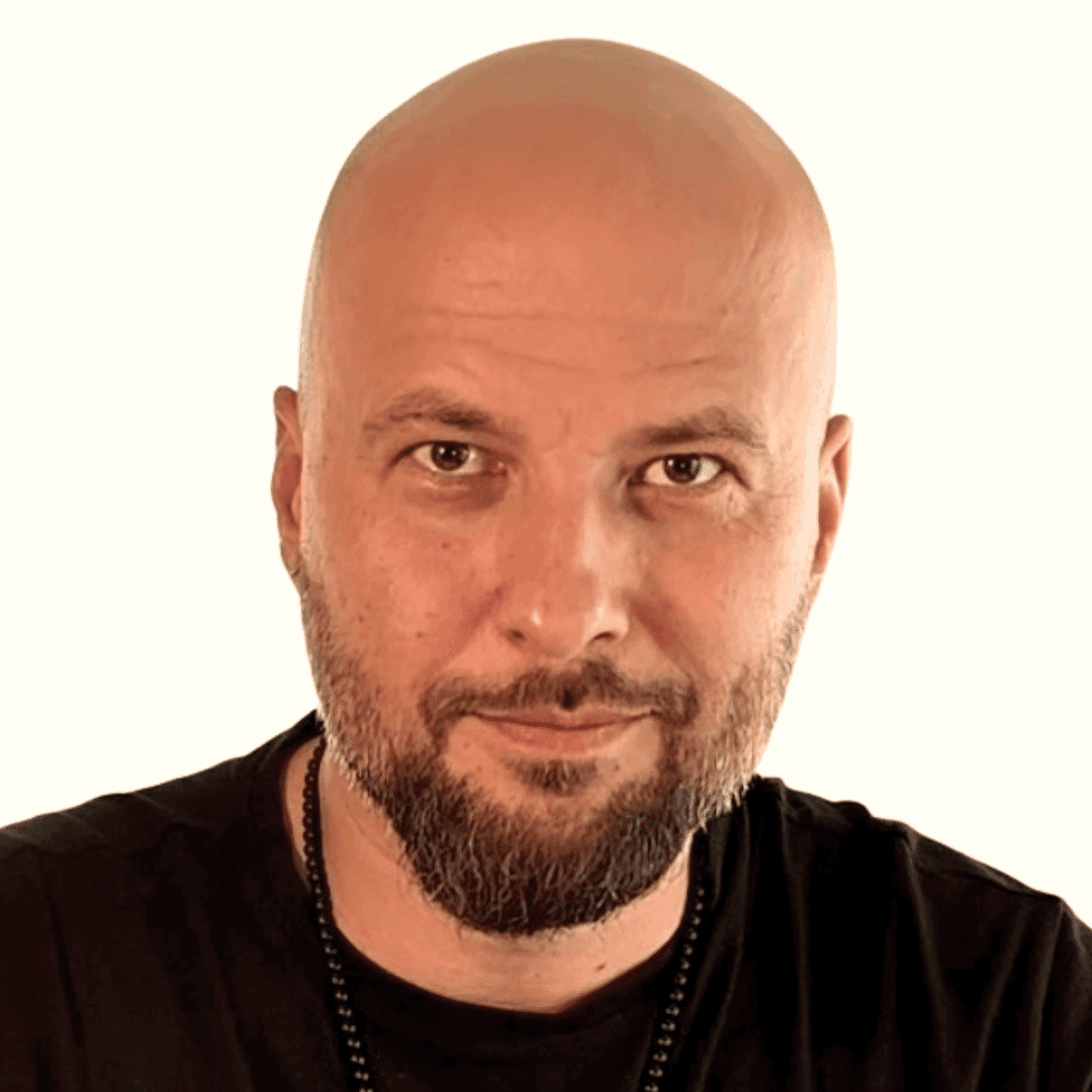 A bald man with a trimmed beard and mustache, wearing a black shirt and a beaded necklace, looks directly at the camera with a slight smile against a plain light background, embodying the calm confidence of the Brain Heart Way.