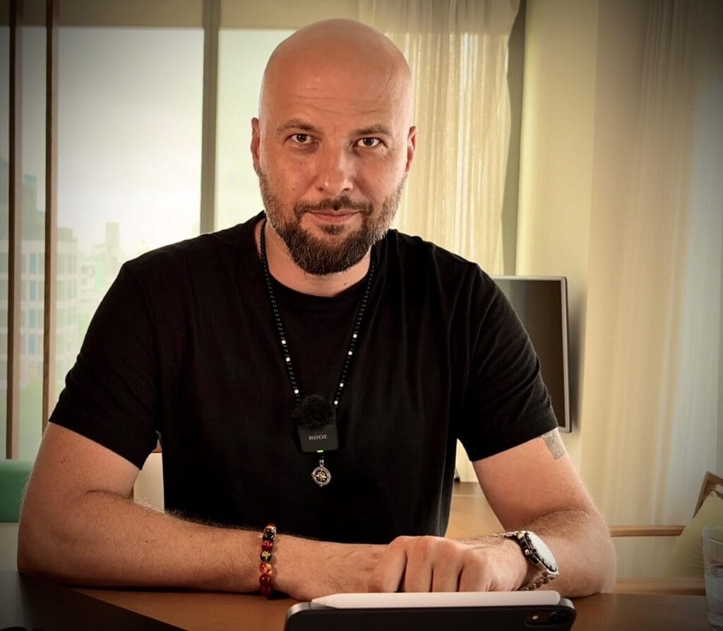 A bald man with a beard, wearing a black shirt and beaded necklace, sits at a desk with a tablet in front of him. Indoors with large windows behind him, he looks directly at the camera, embodying the calm focus of Spiritual Healing - Life Script Hacker